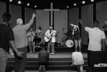 A black-and-white photo of a group standing in a church, listening to someone at the front playing a guitar