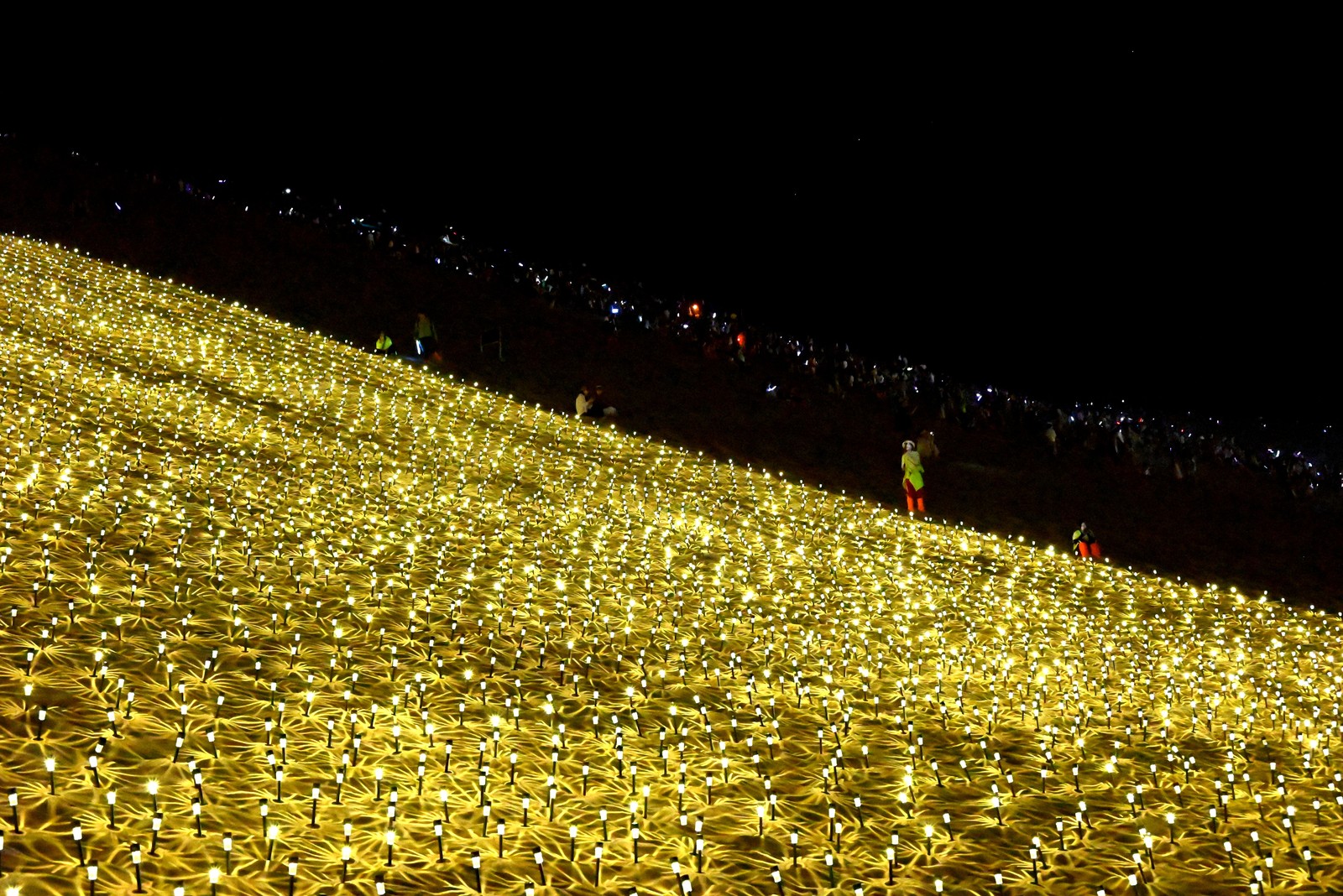 People walk on a mountain slope, past thousands of small lights arranged on the ground.