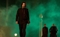 Keanu Reeves, as John Wick, walks toward the camera, green smoke billowing around him and a stoic expression on his face.