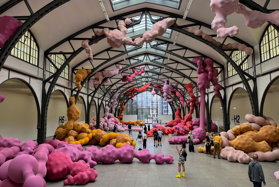 People stand in a large exhibition space looking at artwork that appears to be made of varying sizes of balls stuffed into long colored tubes of stretchy fabric.