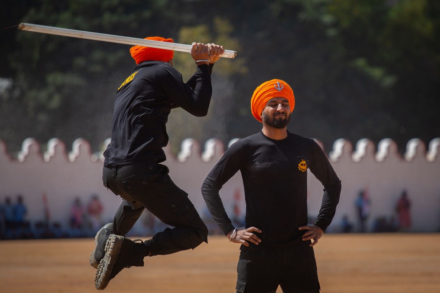 One person leaps in the air, swinging a long glass tube, about to strike another person who stands waiting, hands on hips.