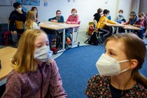Two young girls with masks on are near the camera, and more children with masks can be seen at tables in the background