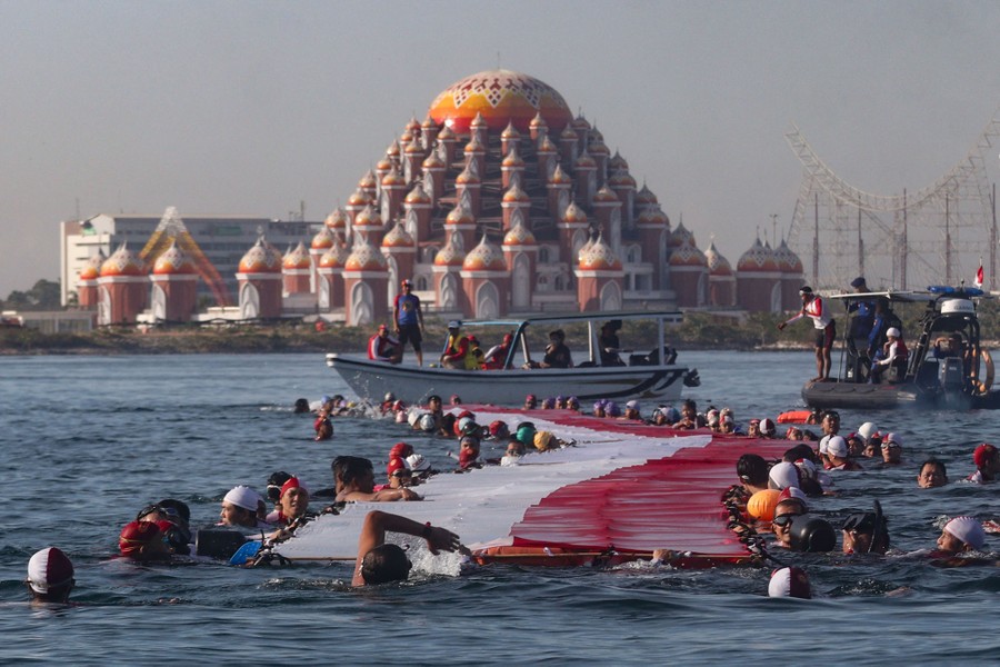 Dozens of people swim, holding on to a long floating sheet of red-and-white fabric.