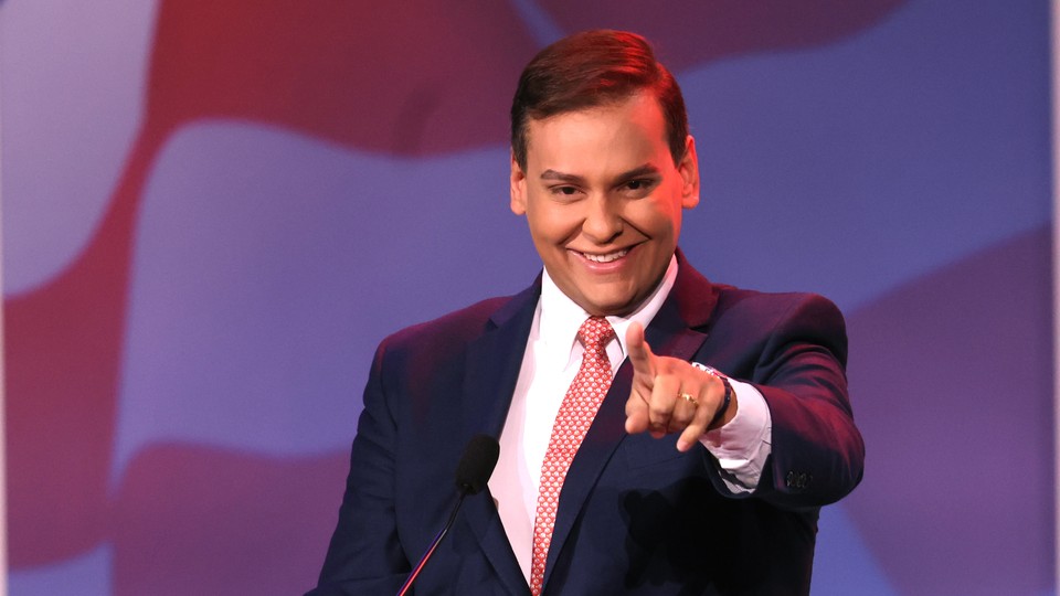 George Santos stands at a lectern and points toward the camera, smiling.