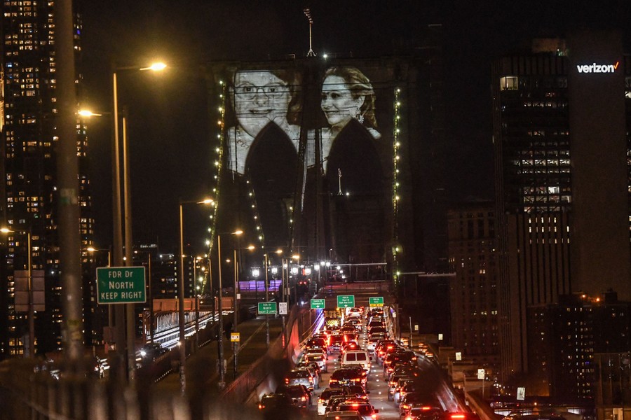The images of faces are seen projected on the side of bridge supports at night, among city buildings and traffic.