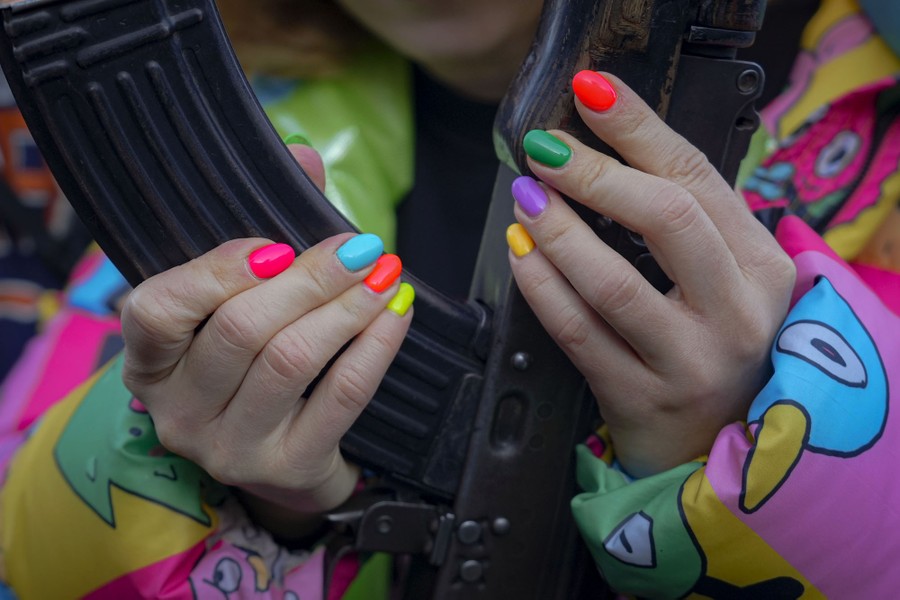 A person wearing a brightly-colored coat and with multi-colored fingernails, handles a rifle and its ammo clip.