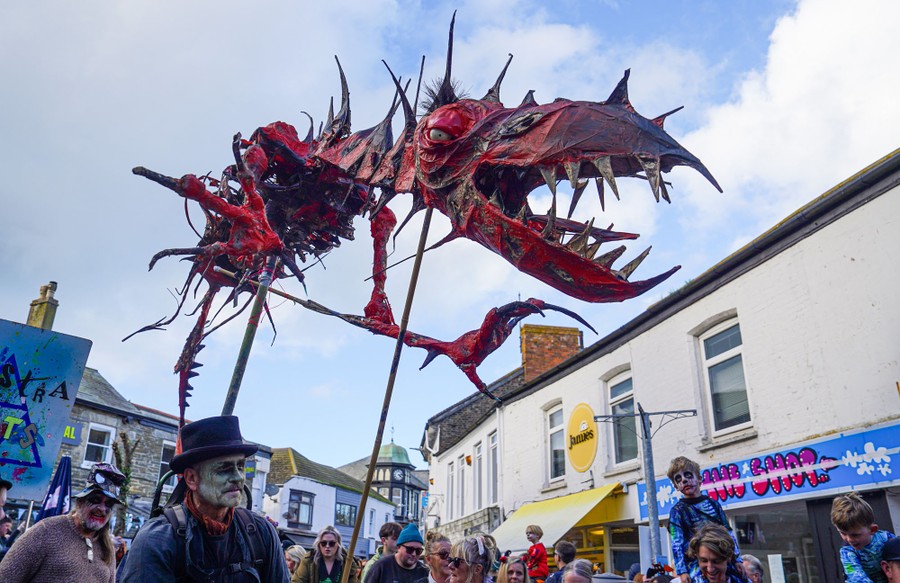 People in costume parade through a street while carrying a large, frightening dragon puppet.