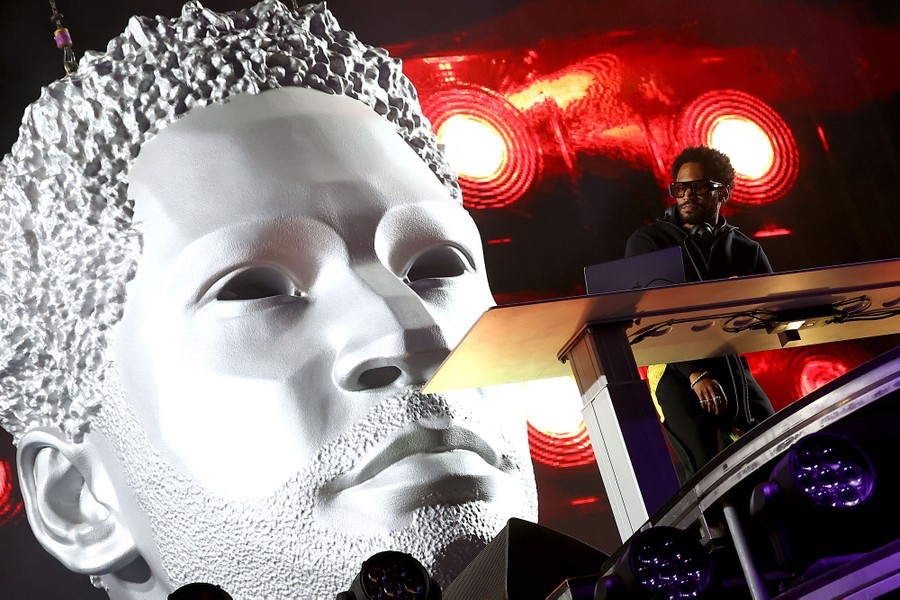 A performer stands onstage in front of a giant sculpture of his head.