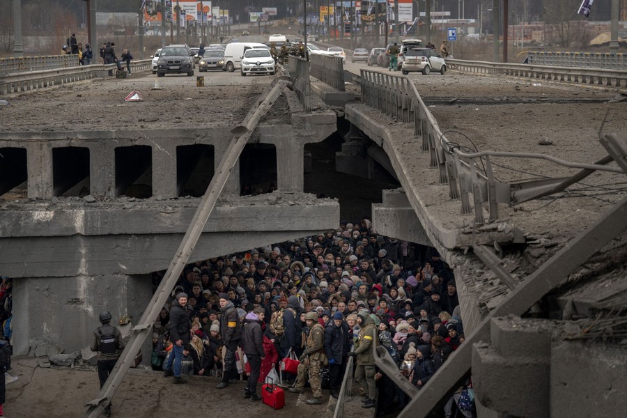 A large crowd of civilians mass under part of a destroyed road bridge.