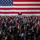Donald Trump addresses a crowd of supporters with a large American flag behind him.