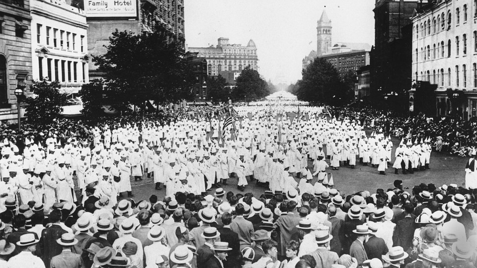 A large crowd of people wearing white gowns and white, pointed hats approaches a fence, against which stand other people in regular clothes.