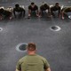 A line of men in green clothing do push-ups on a gray floor
