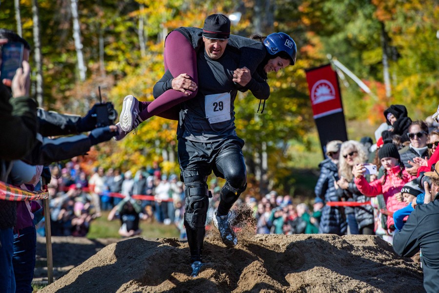 A man carries a woman on his shoulders while running a race, watched by many spectators.