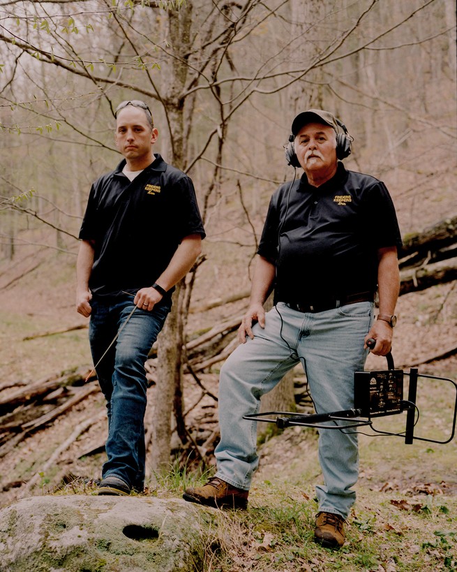 Photo of two men wearing black logo shirts and jeans standing in woods, one with headphones and holding metal detector