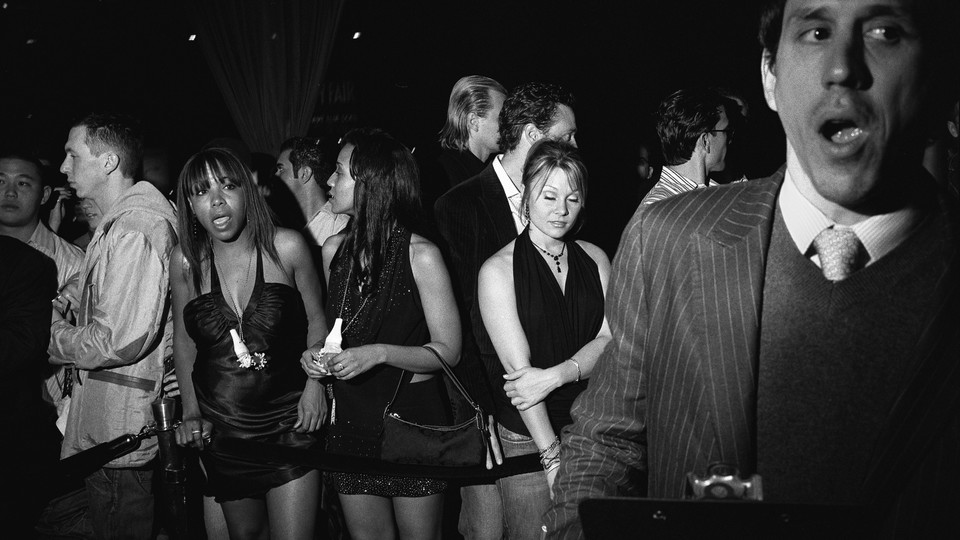 Black-and-white photo of people lining up in front of a nightclub next to a man in a striped suit holding a clipboard