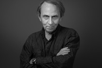 A thin man in a denim shirt poses with his arms crossed in a black-and-white photo.