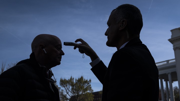 A journalist having his temperature checked by a White House staffer, both shrouded in shadow