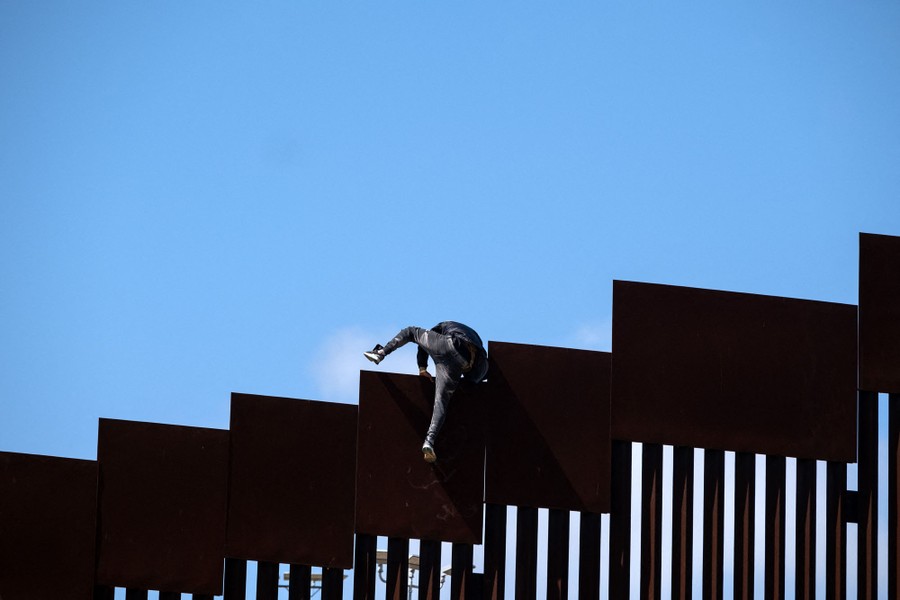 A person climbs over the top of a tall border fence.