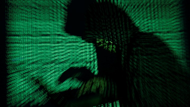 Someone in a hoodie works at a computer, with the background covered in black and green computer code.