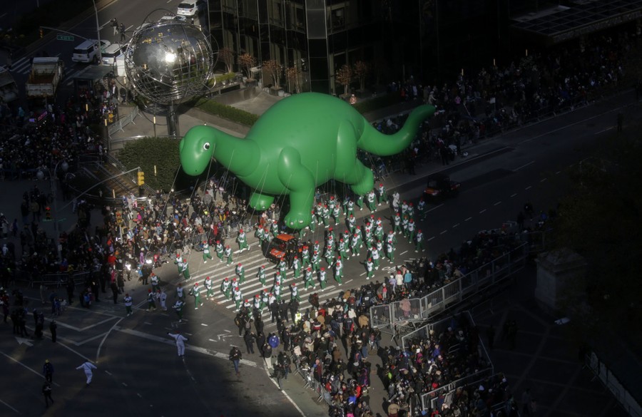 A large green dinosaur balloon floats above a parade route in New York.