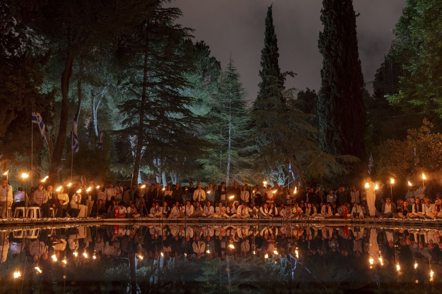 People sit and stand together, holding torches, along a still pool of water.