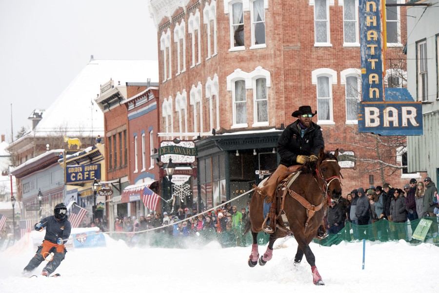 A rider on a horse uses a rope to tow a skier down a snow-covered city street.