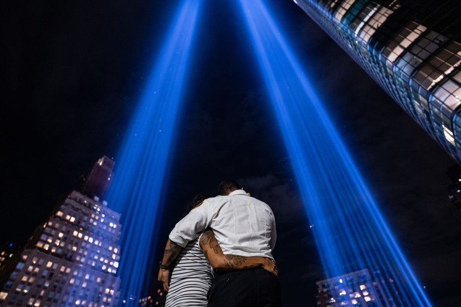 Two people embrace while standing beneath twin pillars of light stretching into the night sky above them and the city.