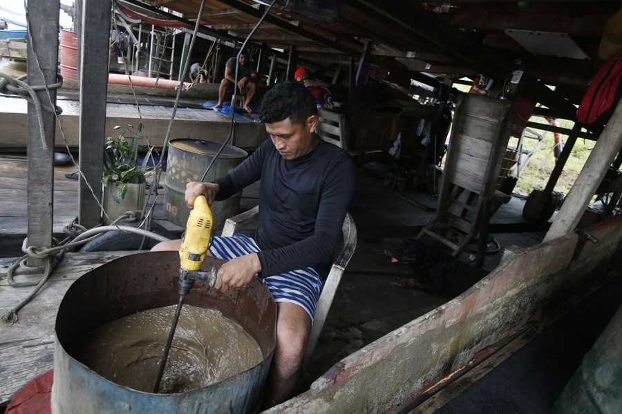 A person uses a large power drill to stir river water inside an open barrel.