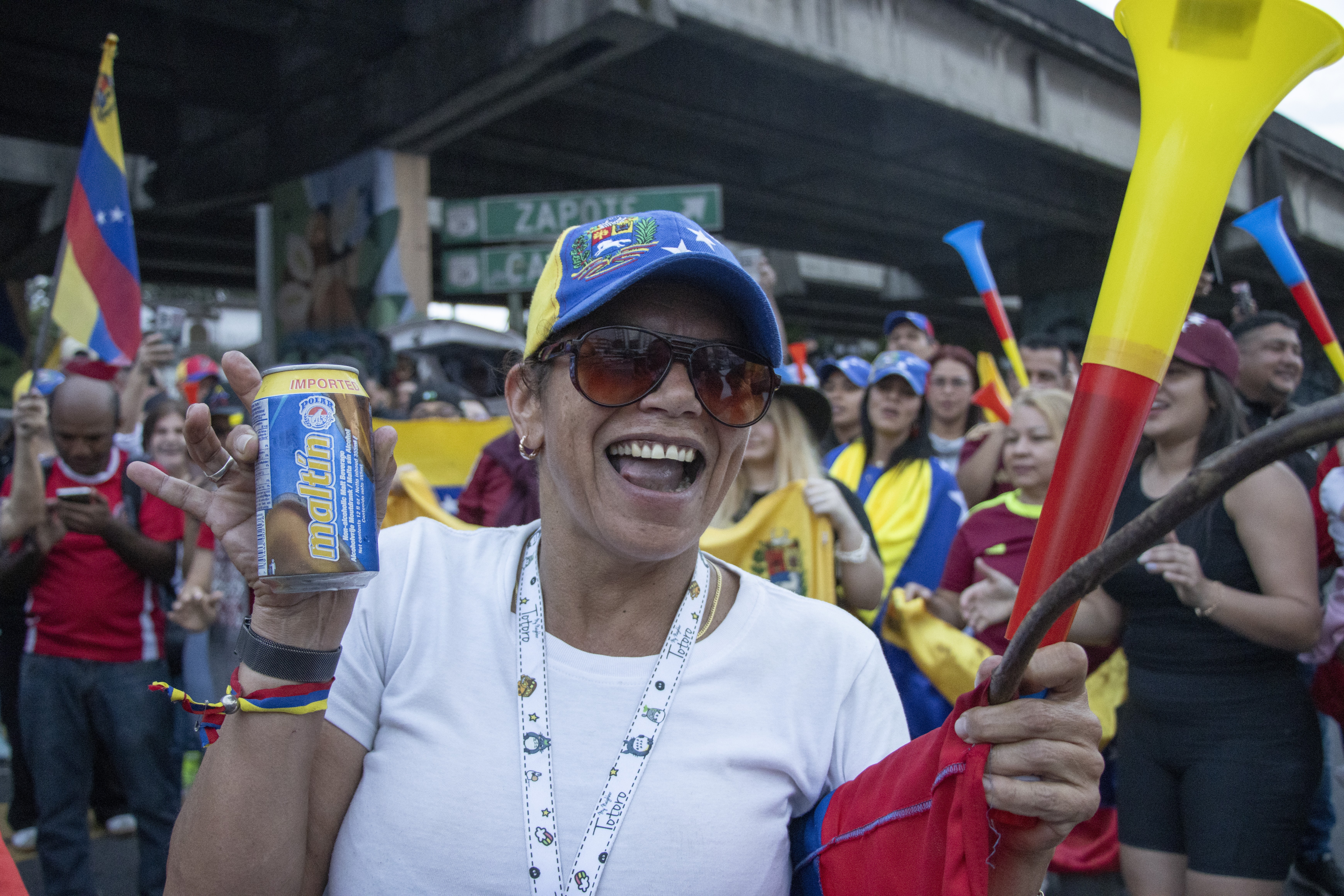 A person smiles and celebrates, holding up a noisemaker and a canned drink.