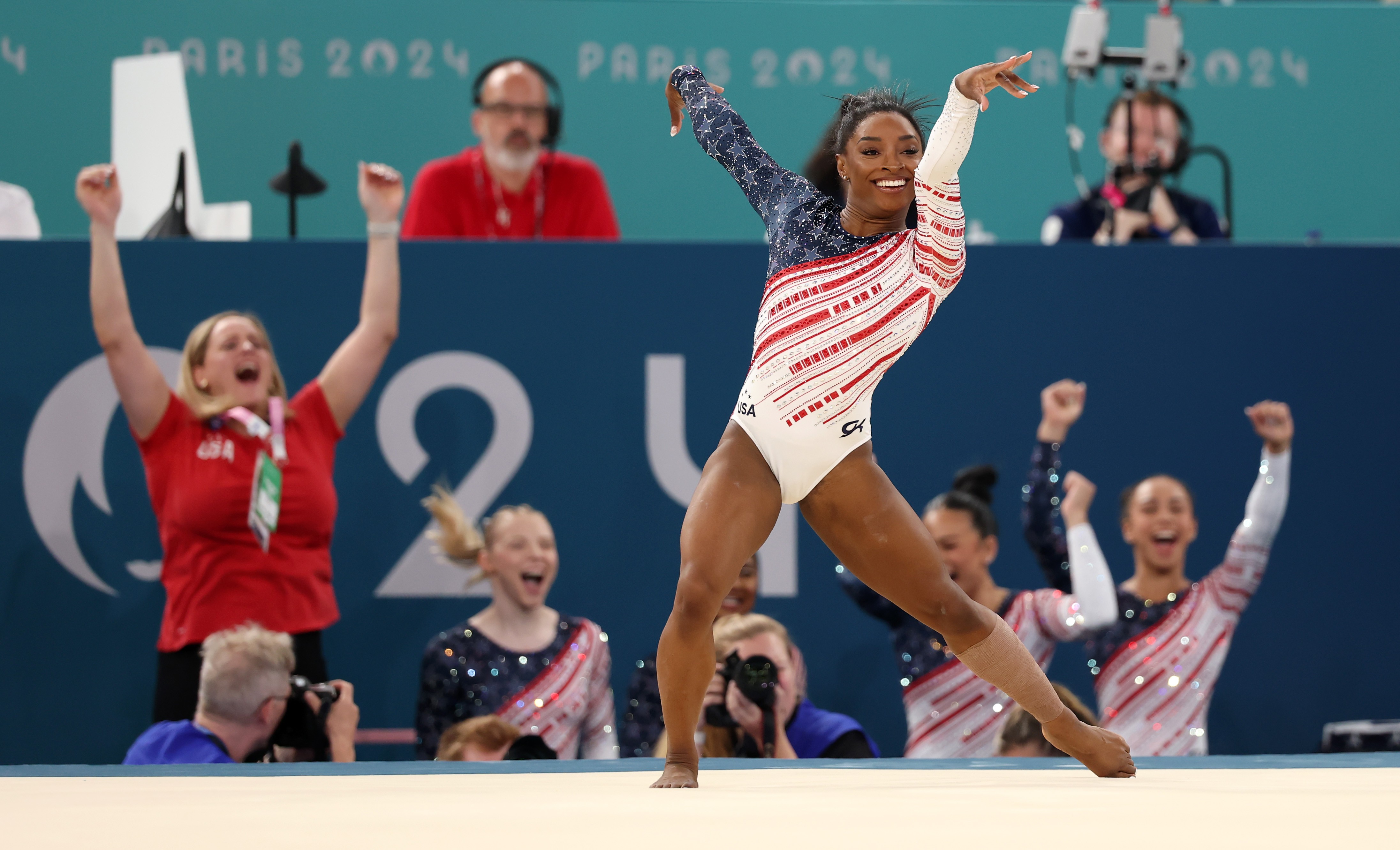 Teammates cheer in the background as a gymnast complete a floor exercise routine.