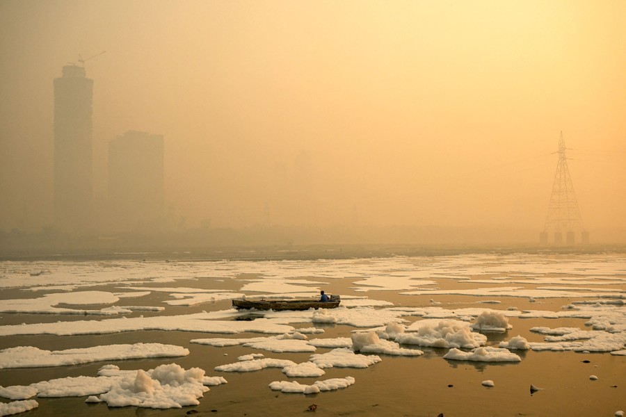 A person steers a small boat through a polluted river beneath a smoggy sky.