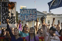 Supporters of the Standing Rock Sioux have signs reading “Oil, Coal, Gas = Climate Chaos,” and “Water is Life.” Their fists are raised, with the White House in the background.