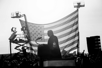 Black-and-white photo of Donald Trump giving a speech to a crowd in front of a large American flag suspended from two bucket lifts