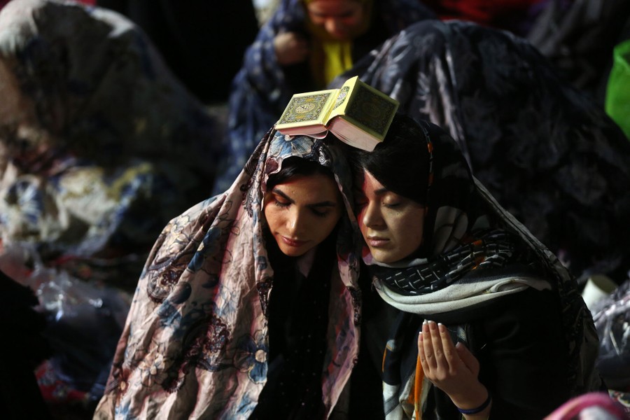 Two Muslim women pray together, a copy of the Quran resting on their touching heads.