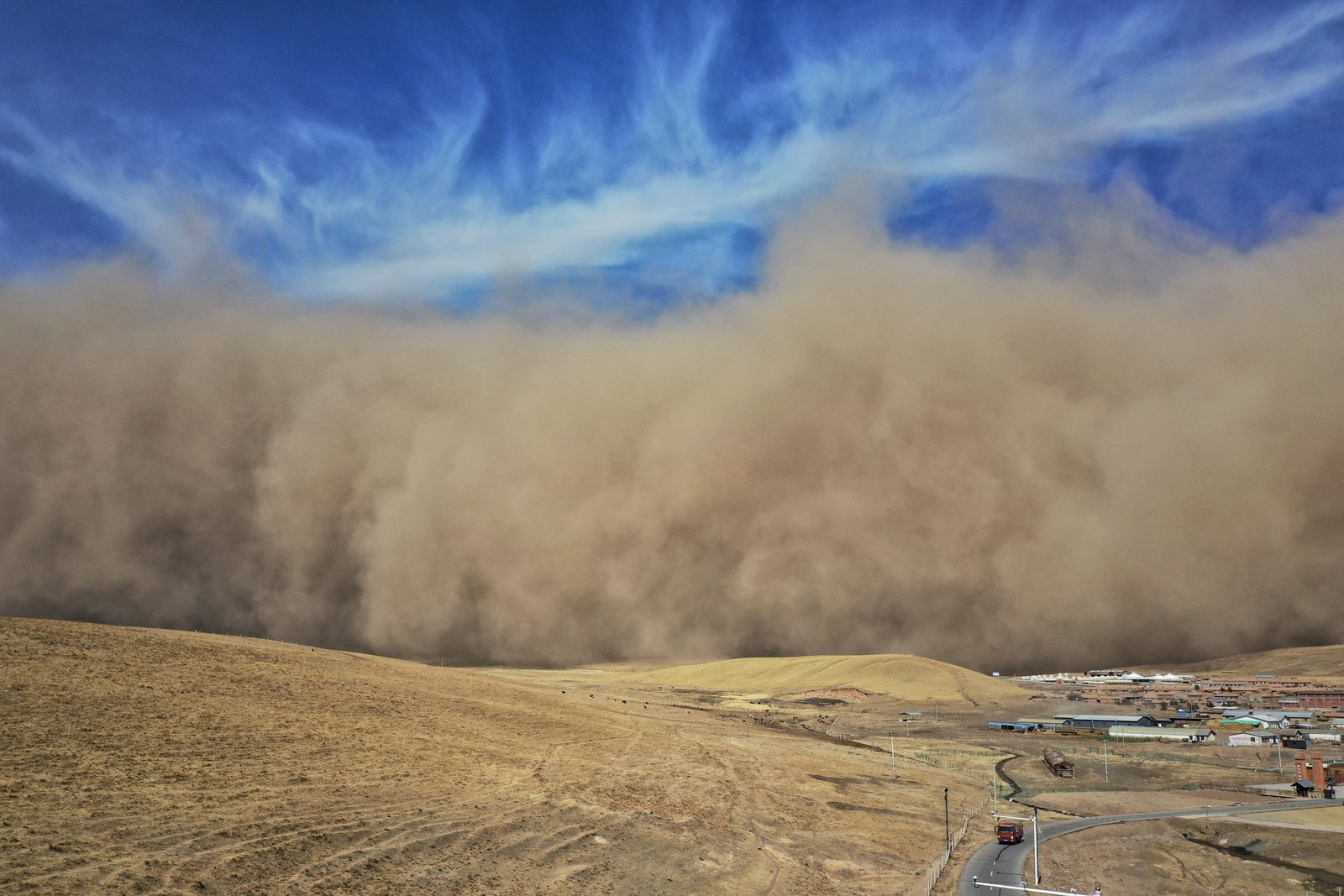 A large cloud of wind-blown sand rolls across the landscape toward a small town, beneath a partly-cloudy blue sky.