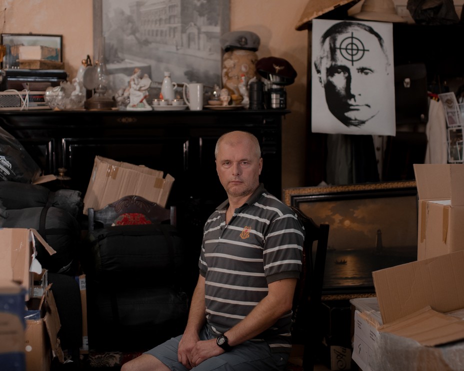 a man sits surrounded by boxes and a Putin image with a target behind him.