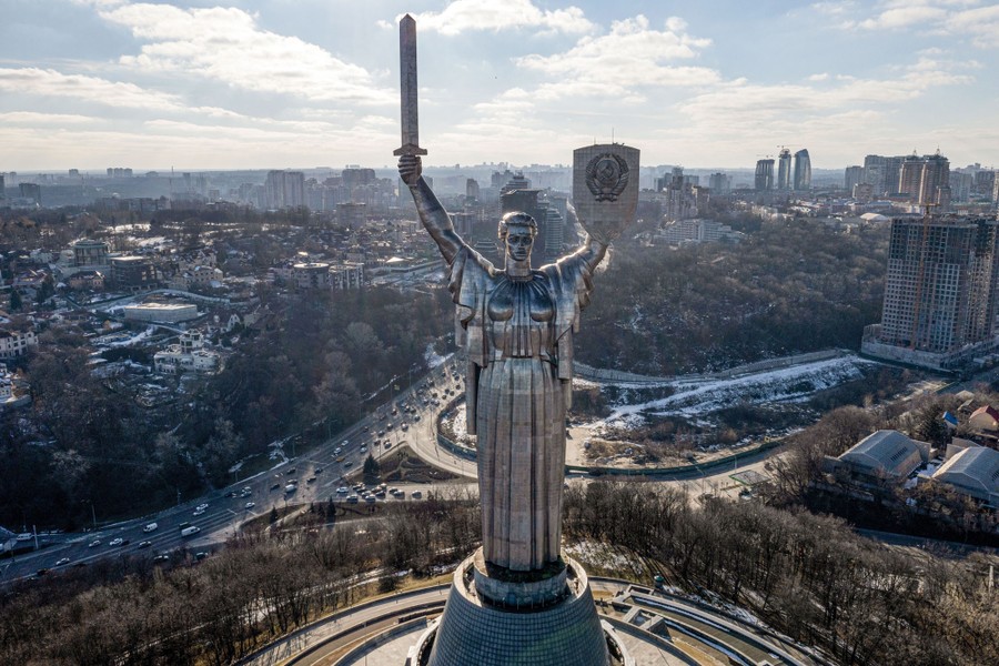 A tall metal-clad statue of a warrior woman holding a sword and shield aloft is seen in front of city buildings.