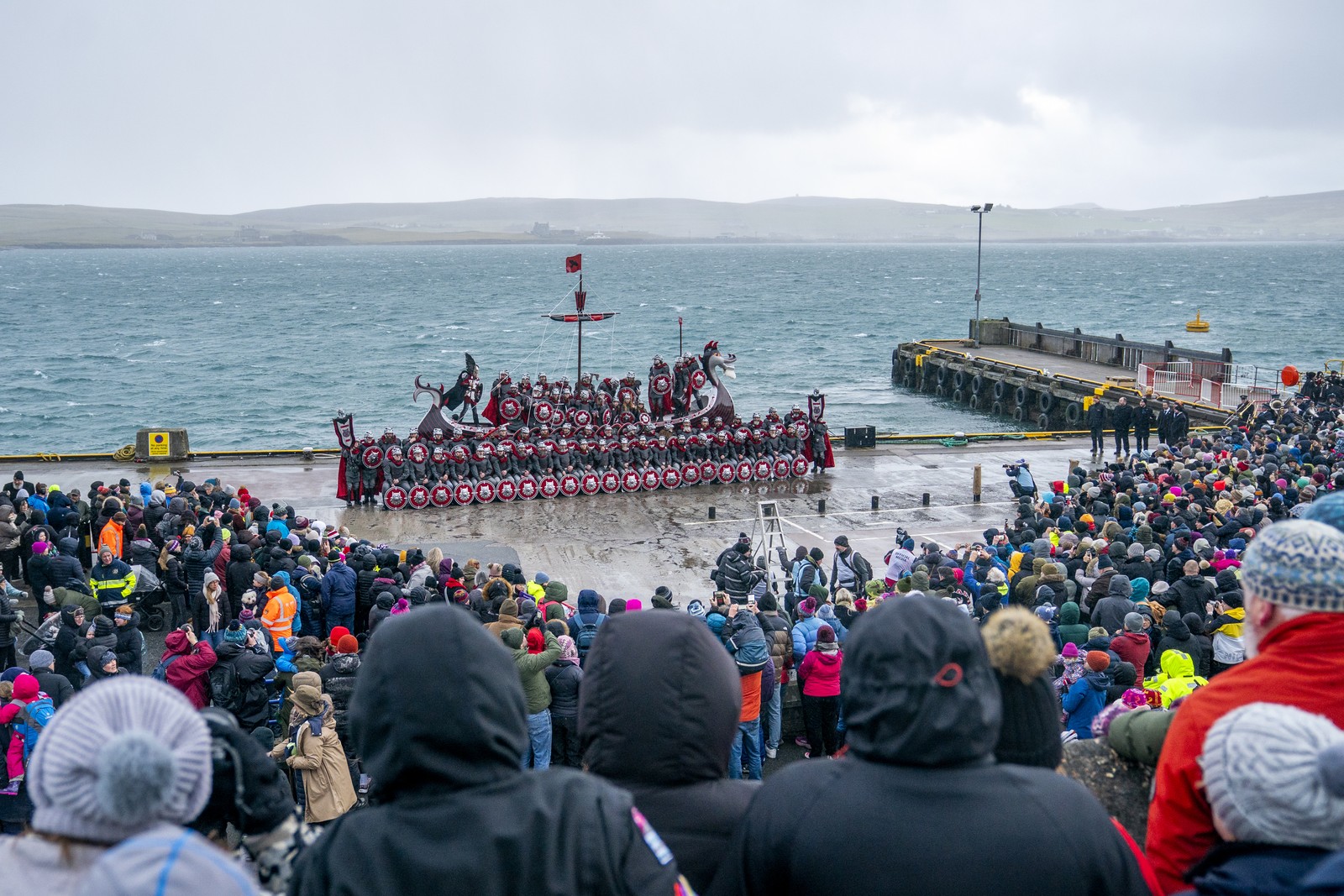 A crowd gathers around a group of people dressed as Vikings who pose beside a replica of a Viking ship beside a harbor.