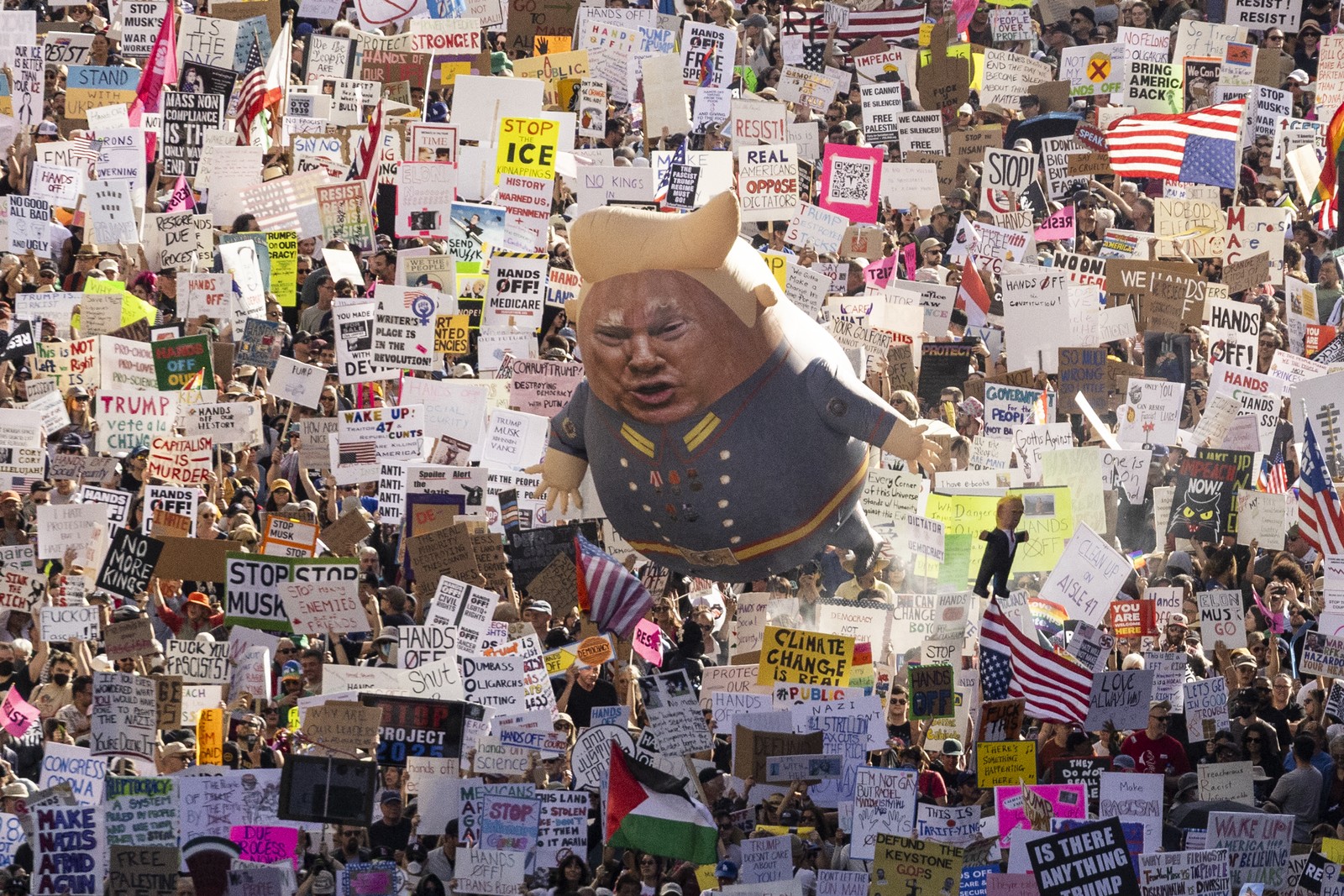 A large crowd of protesters marches down a Los Angeles street carrying many signs and a balloon with an image of President Donald Trump.