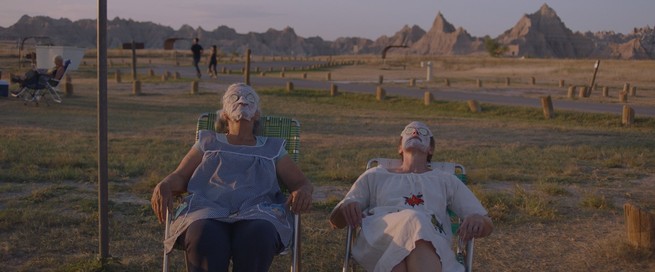 A still from "Nomadland" of two women sitting in lawn chairs in field wearing face masks and with cucumbers on their eyes.