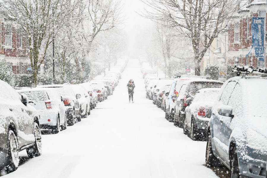 A man walks down the middle of a snow-covered residential street, with cars parked on either side.