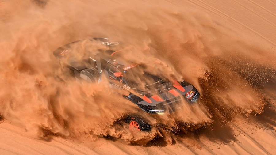 A rally car drives through a desert, kicking up huge clouds of sand and dust.