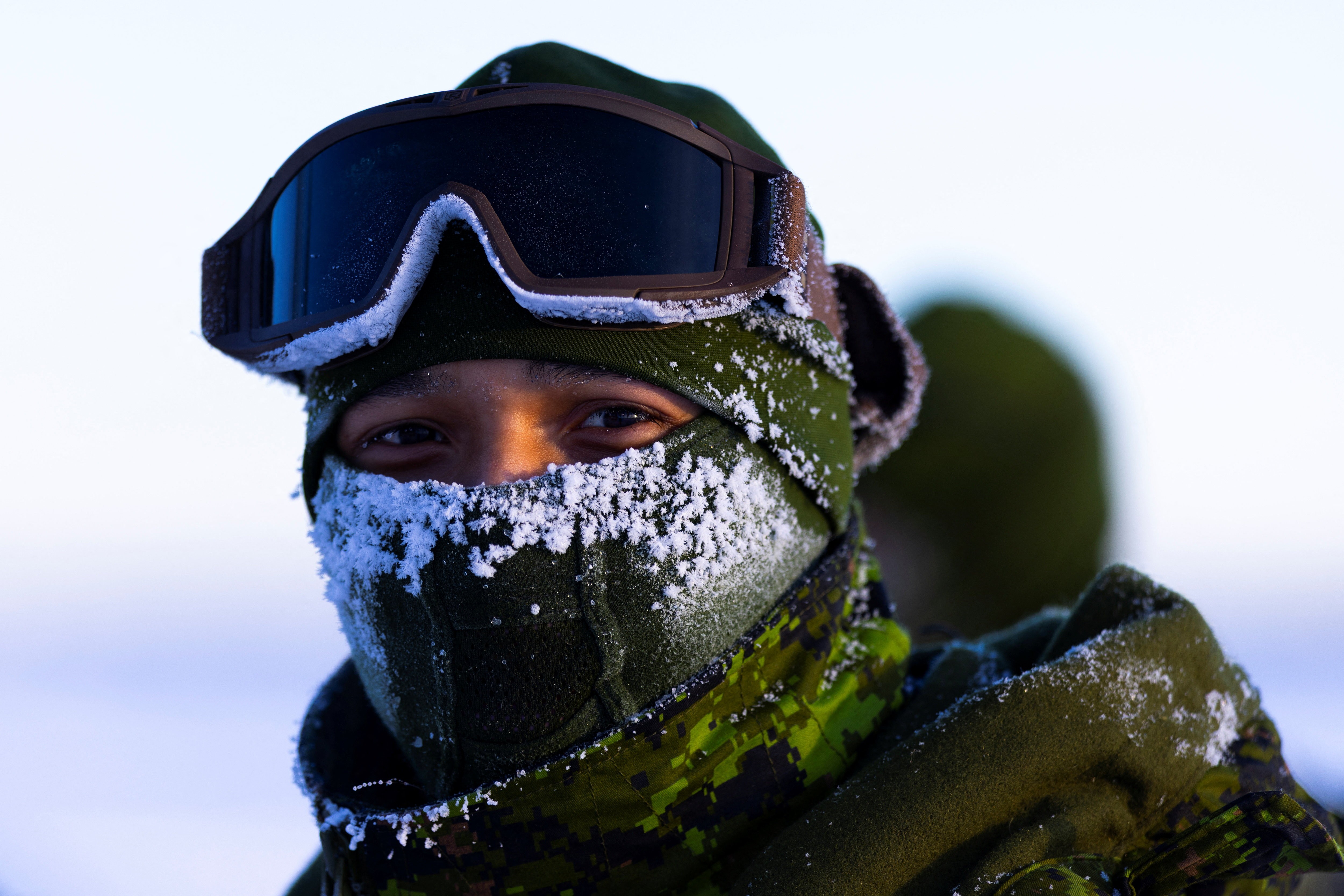 A soldier stands outside on a cold day, with ice buildup around their mask.
