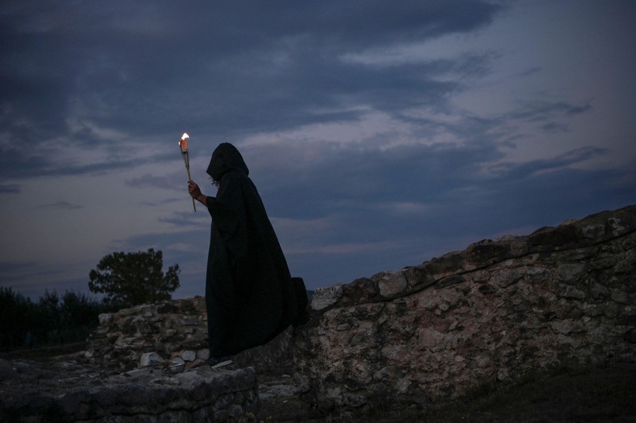 A person wears a dark cloak while carrying a torch and walking on an archeological site.