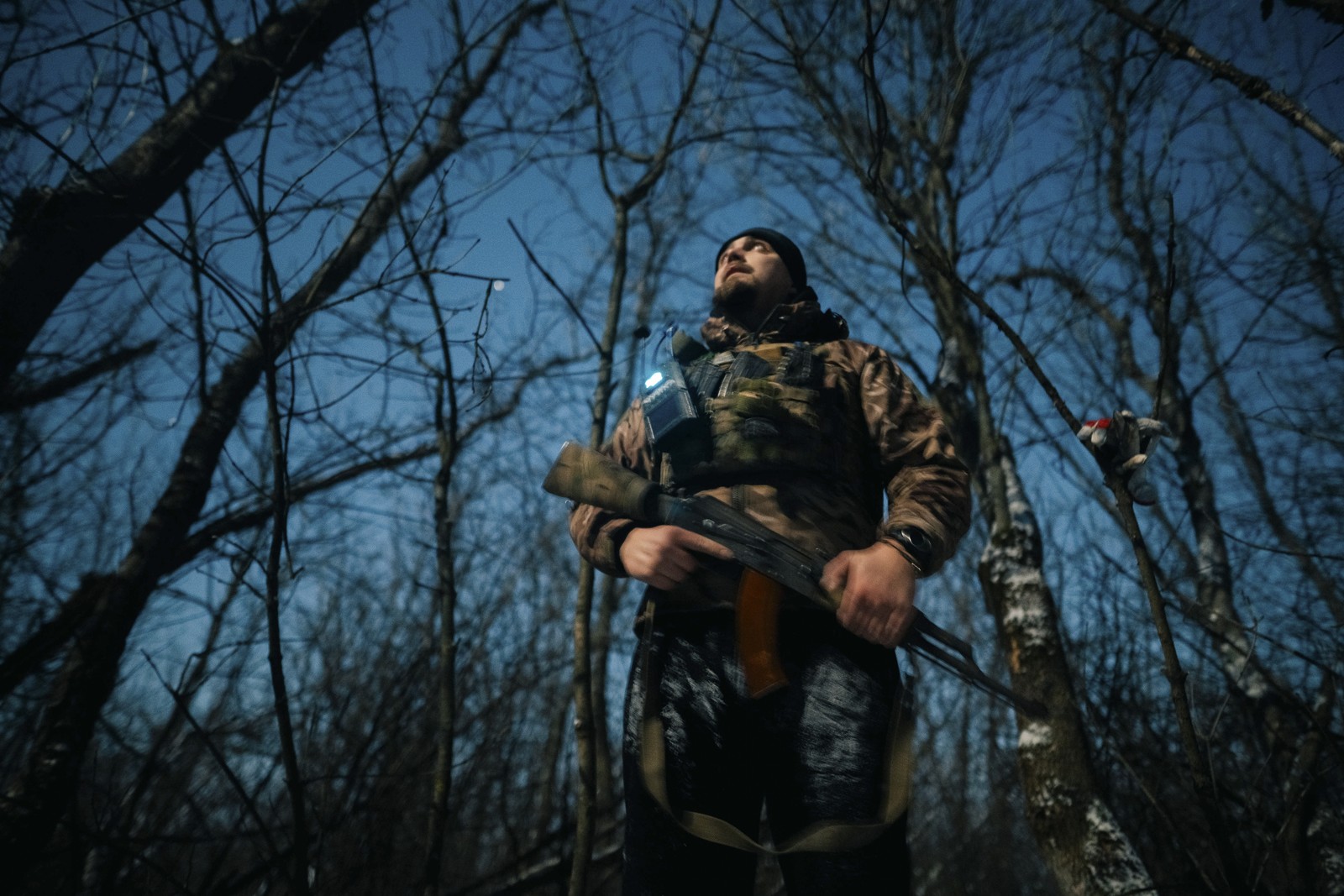 A soldier in a wooded area looks toward the sky.