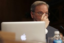 A man sitting behind an Apple laptop covers the lower half of his face with his hand.