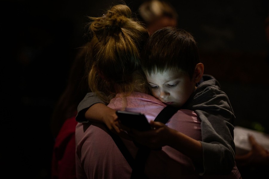 A mother holds her young son in a dark tunnel. His face is lit by the screen of a phone he holds in his hands.