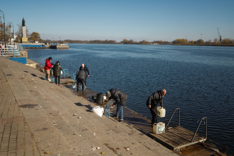 A half-dozen people fill large jugs with water on the steps of a river shoreline.