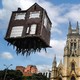 A large art installation, looking like a small, two-story brick dwelling that appears to be floating about 50 feet above a city square, with thick roots hanging beneath the structure.