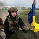 Two members of the Ukrainian 28th Mechanized Brigade ride in an armored vehicle towards hostile Russian forces. The one on the left is smoking a cigarette, the one on the right is scowling, and to their side is a blue and yellow flag.